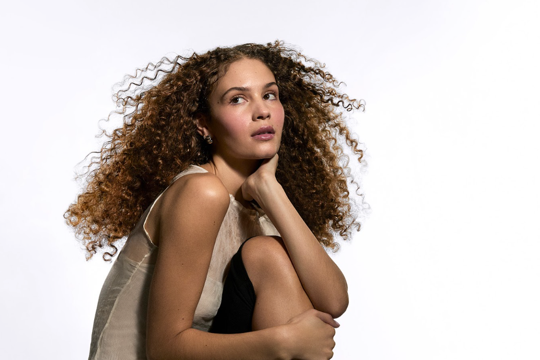 Woman with long, voluminous auburn curls crouching against a white background, resting her chin on her hand