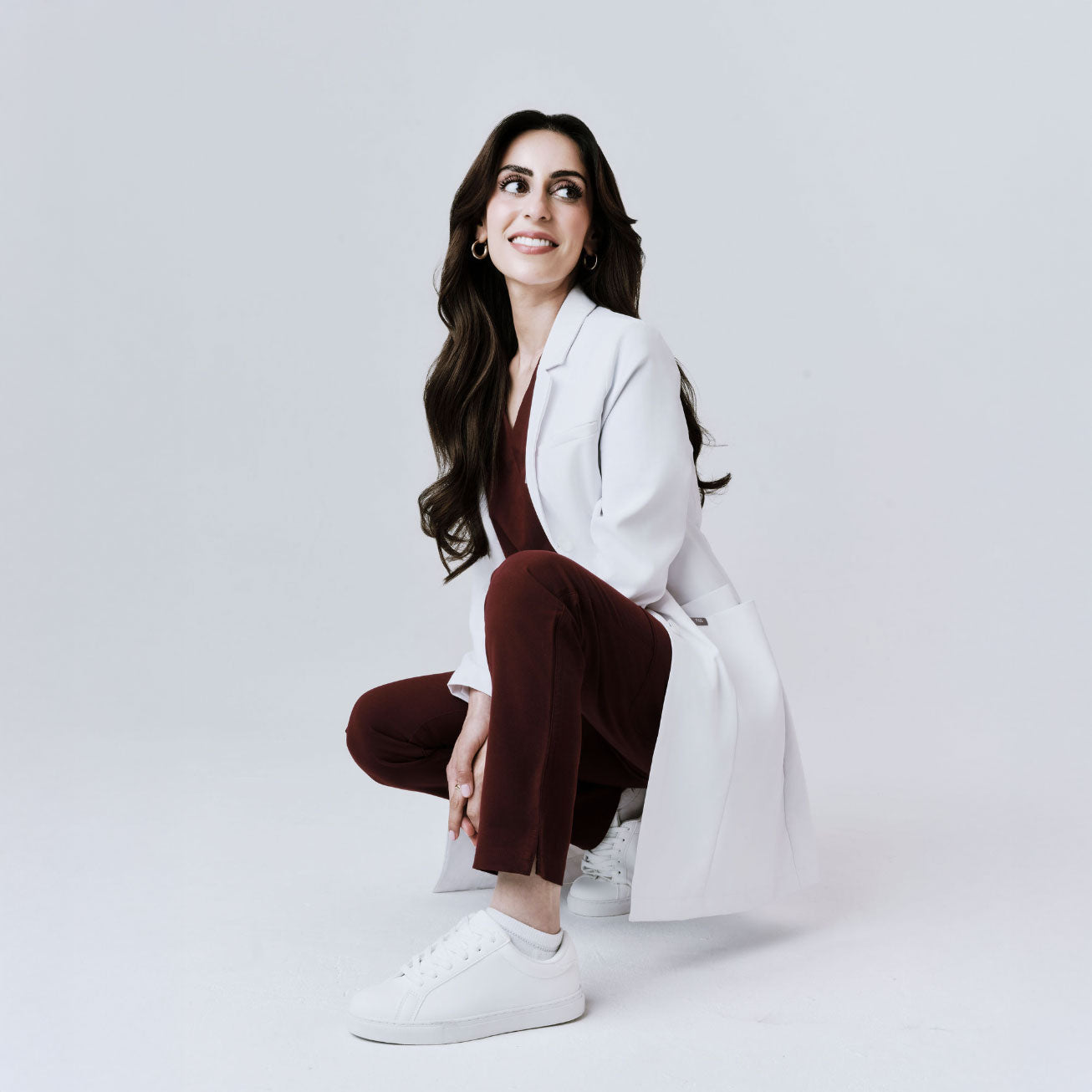 Dr Aamna in a white coat and brown pants sitting on a white background.
