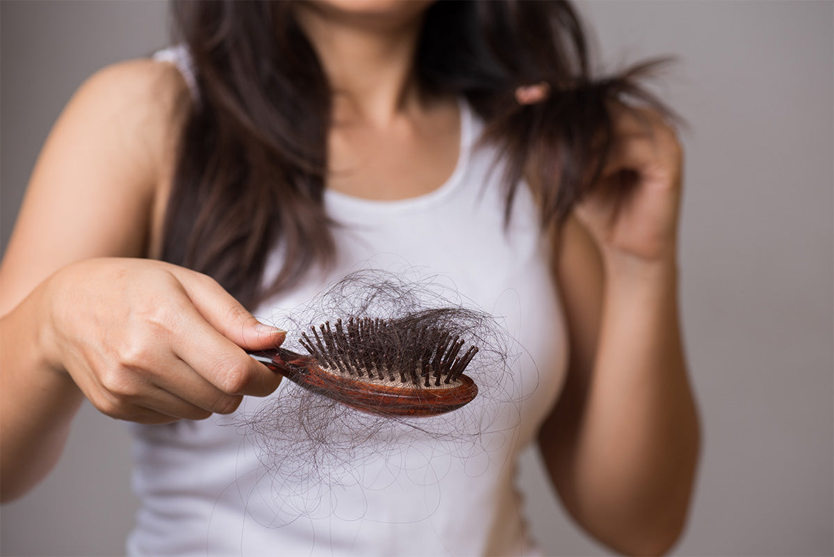 Woman with dark brown hair wearing a white top and holding a hairbrush filled with a hair clump