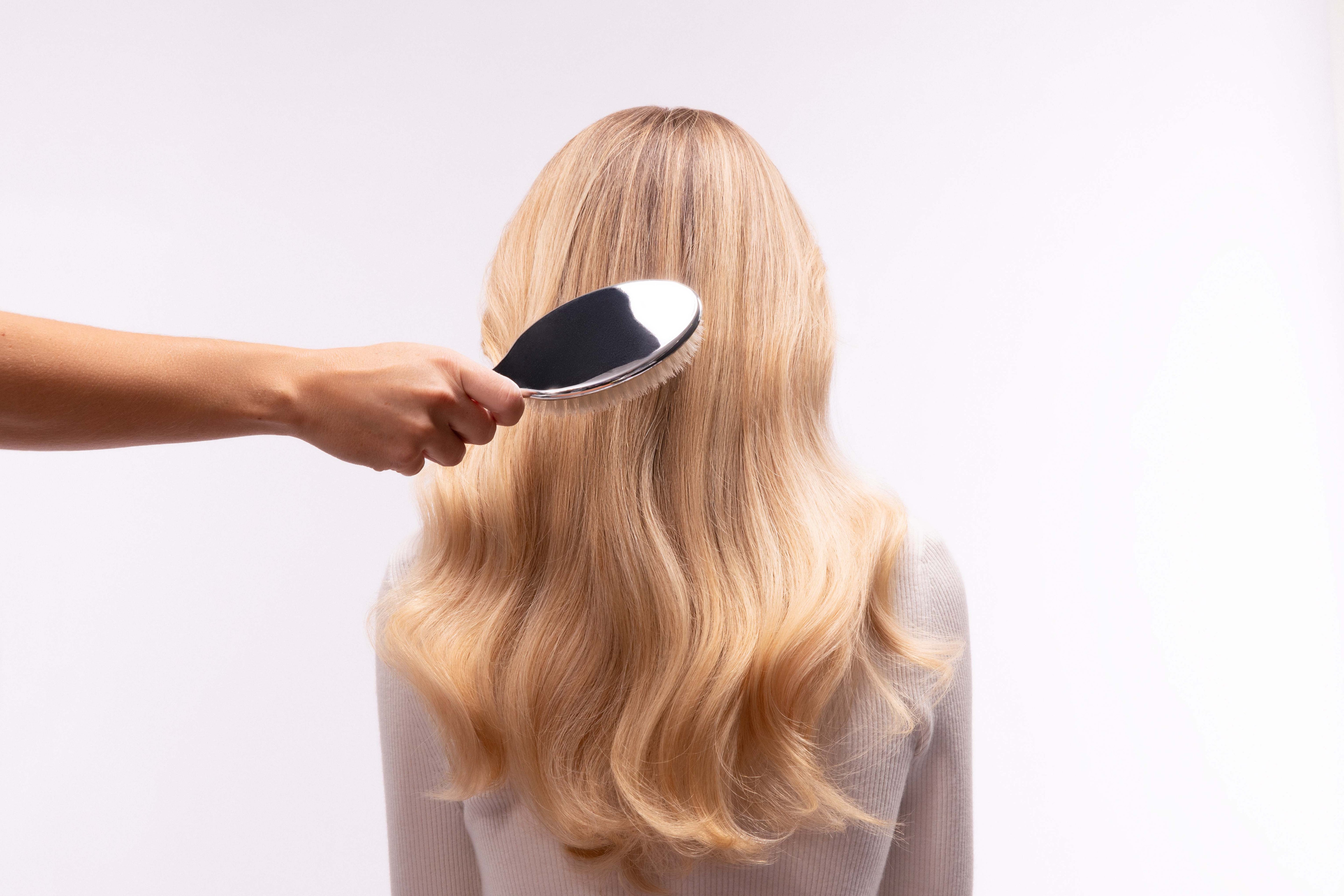Woman with long blonde wavy hair from behind being brushed with a silver paddle hairbrush