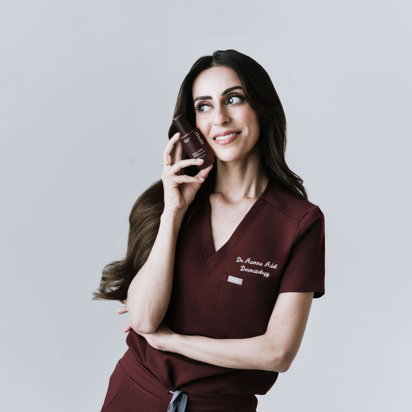 Woman in a maroon scrub top holding hair serum against a light gray background