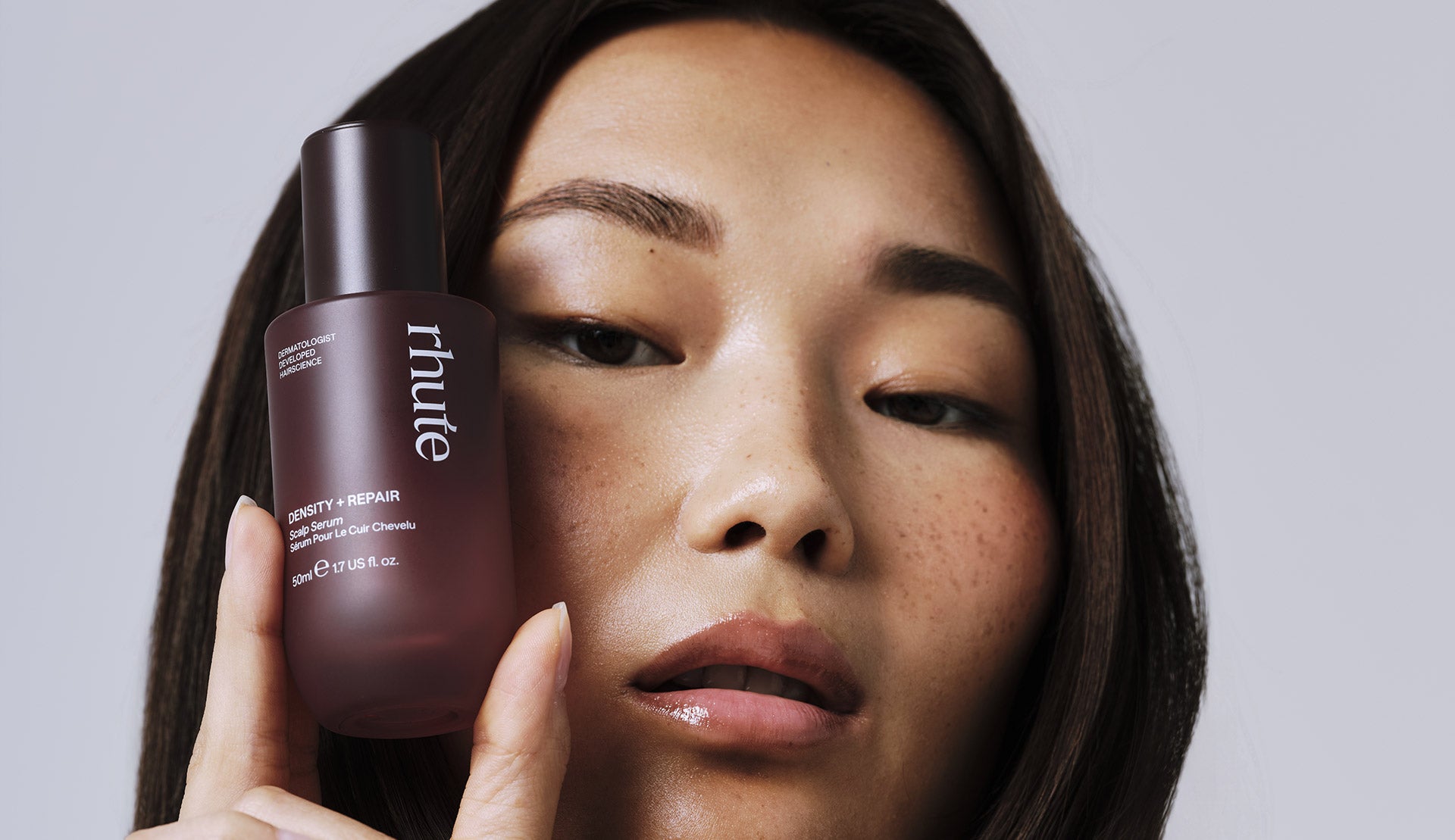 Woman holding a purple skincare bottle with 'rhute' branding against a neutral background