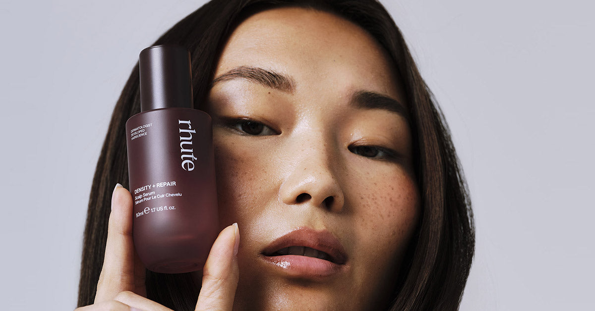 Close-up of a woman holding a frosted maroon bottle of rhute “Density + Repair” scalp serum beside her face.